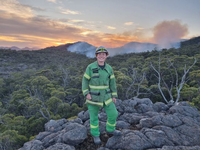 Kayla is wearing her green overall, in the background is smoke from a fire