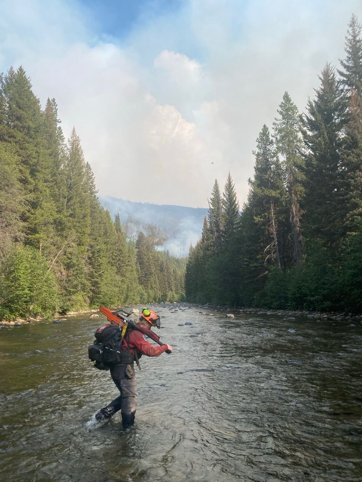 A firefighter carrying a chainsaw crosses a river on foot