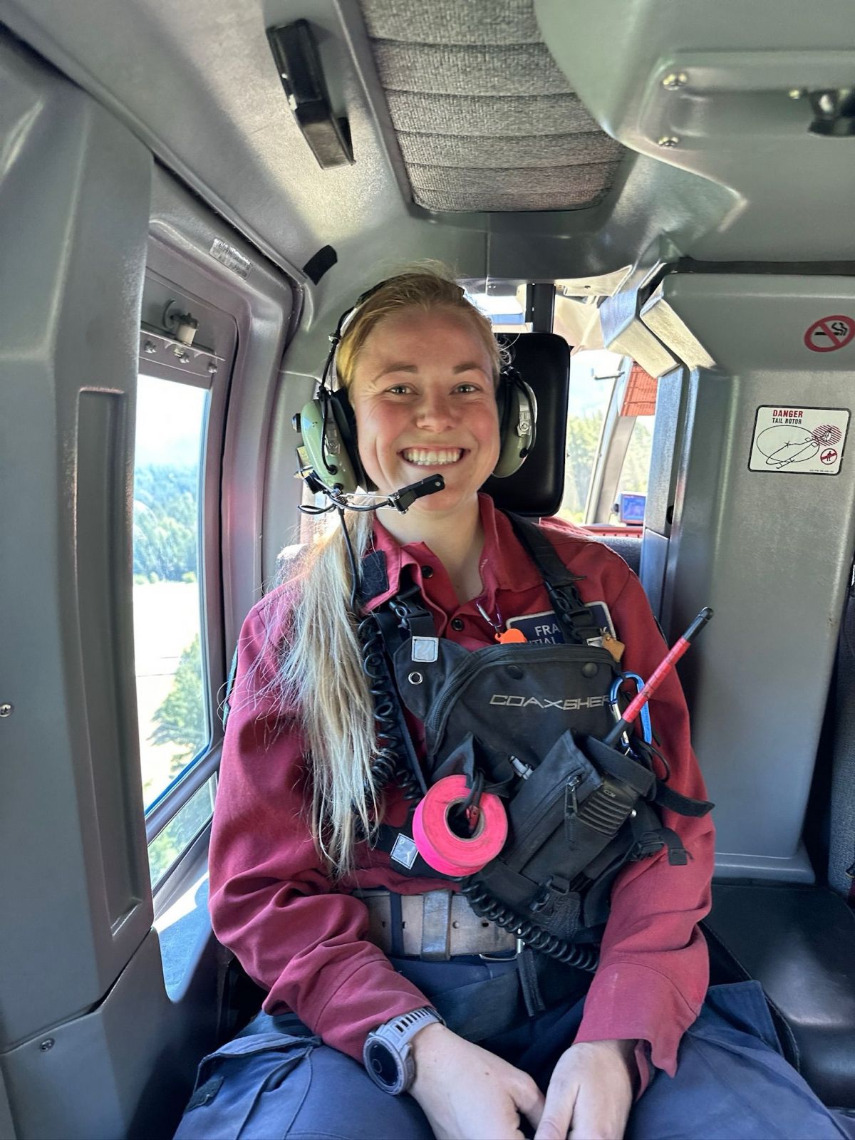 A woman sits in a helicopter with a headset on and smiles at the camera