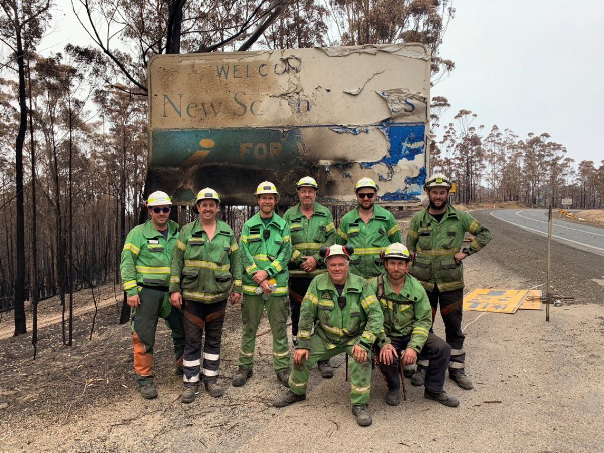 8 Forest Fire Management Victoria (FFMVic) firefighters in front of burnt out Welcome to NSW sign in the fire-affected Mallacoota