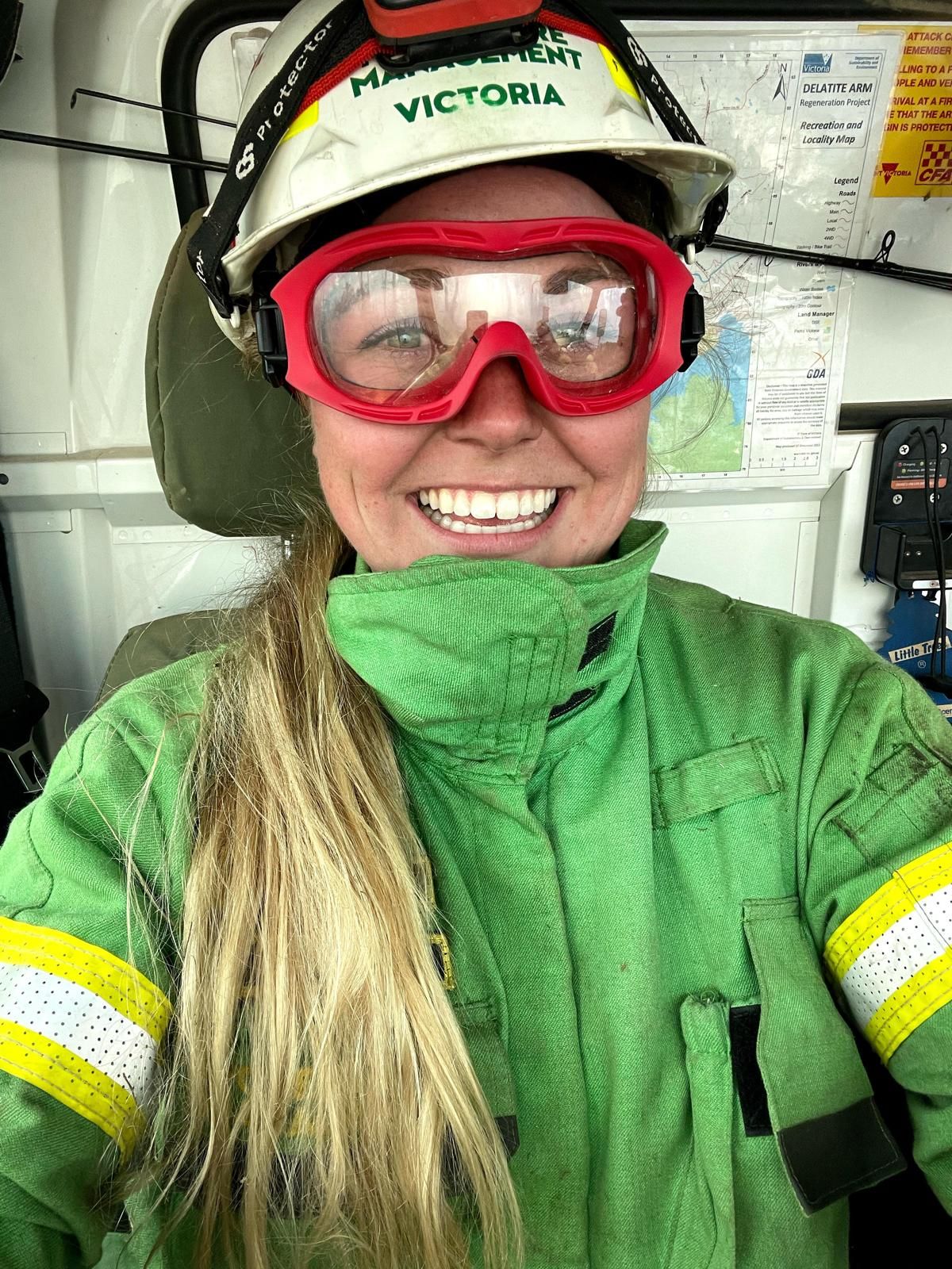 A female firefighter in green uniform and red goggles smiles at the camera