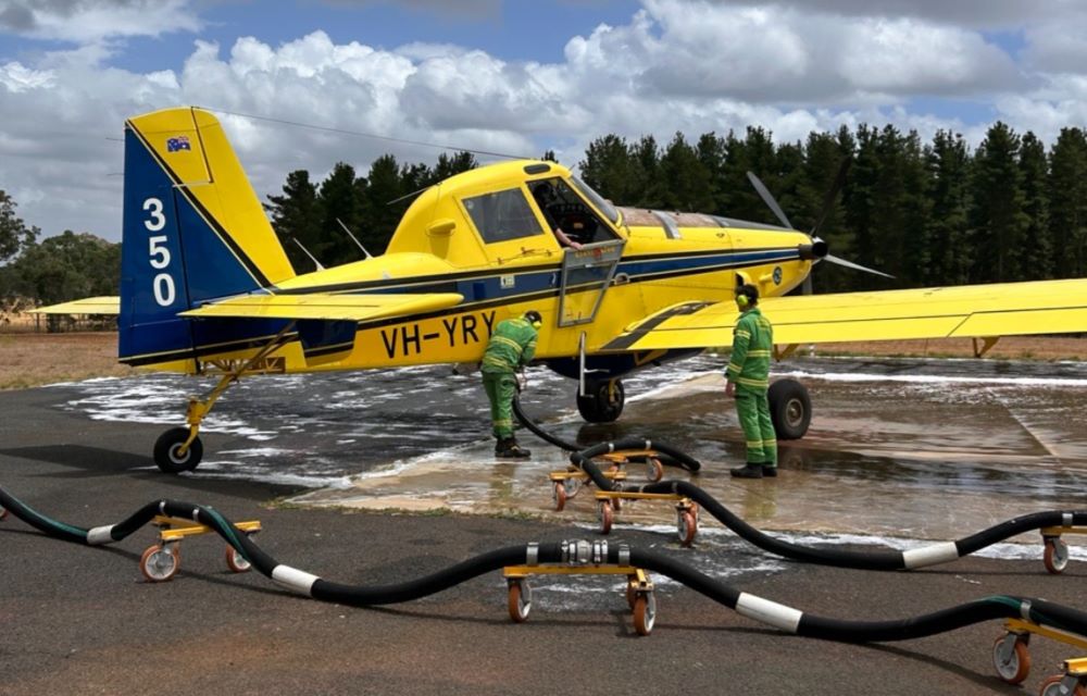 A yellow firefighting aeroplane sits on the tarmac. It is being refuelled by two firefighters in green overalls