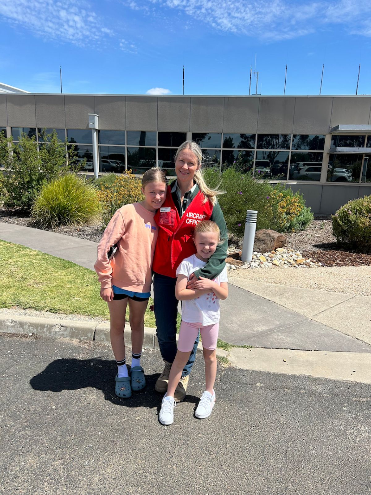 Lauren Todman and their two children stand together in a carpark in front of a low-set building. It is a sunny day with blue skies peering out over the top of the building